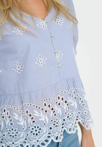 Blouse bleu clair à manches courtes, ornée de broderies florales blanches et d'un ourlet en dentelle festonné. Fermeture à boutons sur le devant.