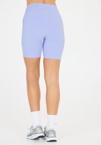 Lila träningsshorts med hög midja, slät textur och ficka på sidan, bärs tillsammans med grå löparskor och vita strumpor.