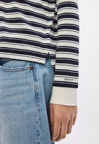 Top cu mâneci lungi, cu dungi albastre și albe, cu deschideri laterale, asortat cu blugi din denim albastru deschis. Logo-ul GANT pe manșetă. Țesătură moale din bumbac.