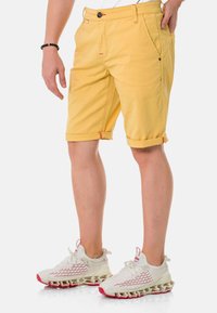 Gelbe Baumwollshorts mit umgeschlagenem Saum, versehen mit roten Akzenten und Fronttaschen. Kombiniert mit leichten weißen Sneakers mit roten Details.