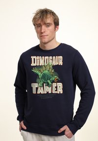 Sudadera azul marino hecha de una tela suave, que presenta un gráfico de dinosaurio y un texto en negrita "DOMADOR DE DINOSAURIOS" en naranja y blanco.