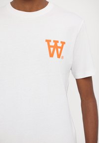 Hvit bomull T-skjorte med en liten oransje grafikk av inverterte bokstaver "W" og "A" på venstre brystområde. Korte ermer, rund hals design.
