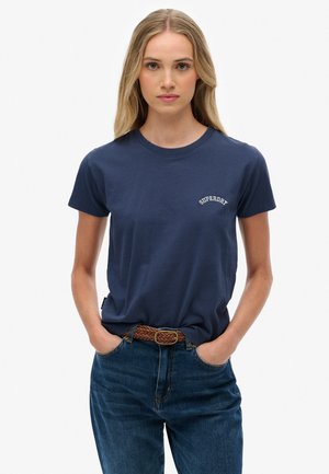 T-shirt en coton bleu marine à manches courtes et col rond, avec un logo blanc sur la poitrine gauche. Porté avec un jean bleu taille haute.