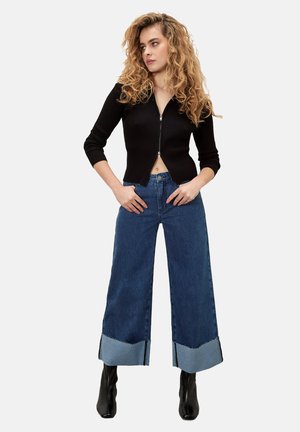 Cardigan court noir à côtes avec fermeture éclair ; jeans en denim bleu à jambes larges avec ourlet contrastant et bottines noires.