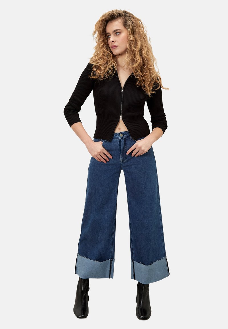 Cardigan nera a costine, cropped, con zip; jeans blue in denim a gamba larga con orlo a contrasto e stivaletti neri.