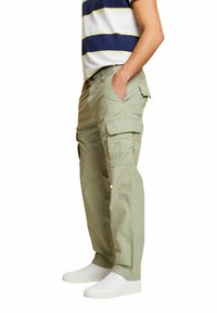 Man in beige cargobroek met meerdere zakken en witte sneakers, staande met handen in de zakken.