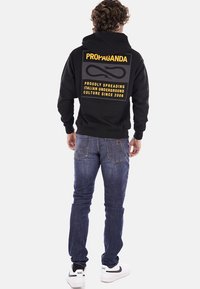 Felpa nera con grande testo giallo sul retro, caratterizzata da un design grafico e una tasca a kenguru. Indossata con jeans blu e scarpe da ginnastica bianche.