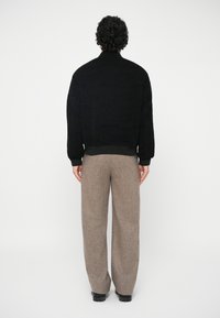 Άνδρας με σπαστά μαλλιά που στέκεται με την πλάτη, φορώντας μαύρο μπουφάν, μπεζ παντελόνια φαρδιά και μαύρα παπούτσια, σε λευκό φόντο.