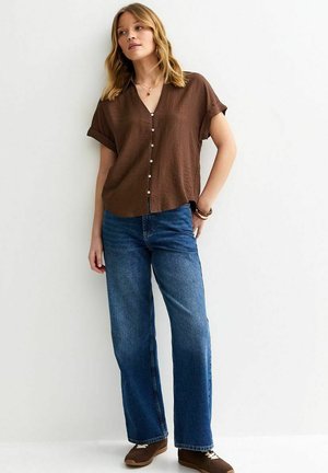 Kobieta ubrana w brązową, krótkorękawową koszulę na guziki, niebieskie szerokie dżinsy i brązowe trampki, stoi na tle białej ściany.