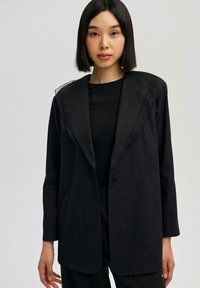 Černý blazer s přízdobou, texturovaným límcem, zapínáním na jeden knoflík a dlouhými rukávy, kombinovaný s černým topem pod ním.