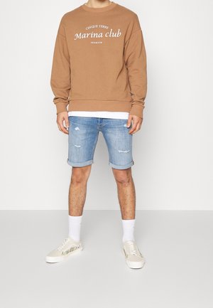 Muž v hnědé mikině s nápisem "Marina club", světle modrých roztrhaných džínových šortkách s ohrnutými nohavicemi, bílých ponožkách a bílých teniskách s květinovým vzorem.