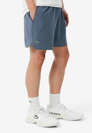 Man draagt blauwe Lacoste-shorts, witte Lacoste-sokken en witte Lacoste-sneakers, staat met handen in de zakken op een witte achtergrond.