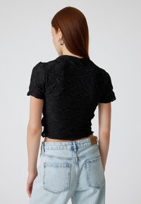 Sort, tekstureret crop top med et mønstret design, korte ærmer og en tætsiddende silhuet, parret med lyseblå højtaljede jeans.