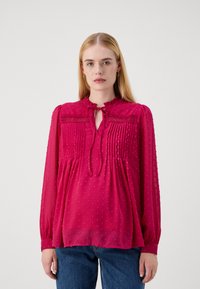 Blusa color lampone con punti testurizzati, dotata di un colletto a legaccio, colletto a volant e parte anteriore plissettata. Maniche lunghe e vestibilità ampia.