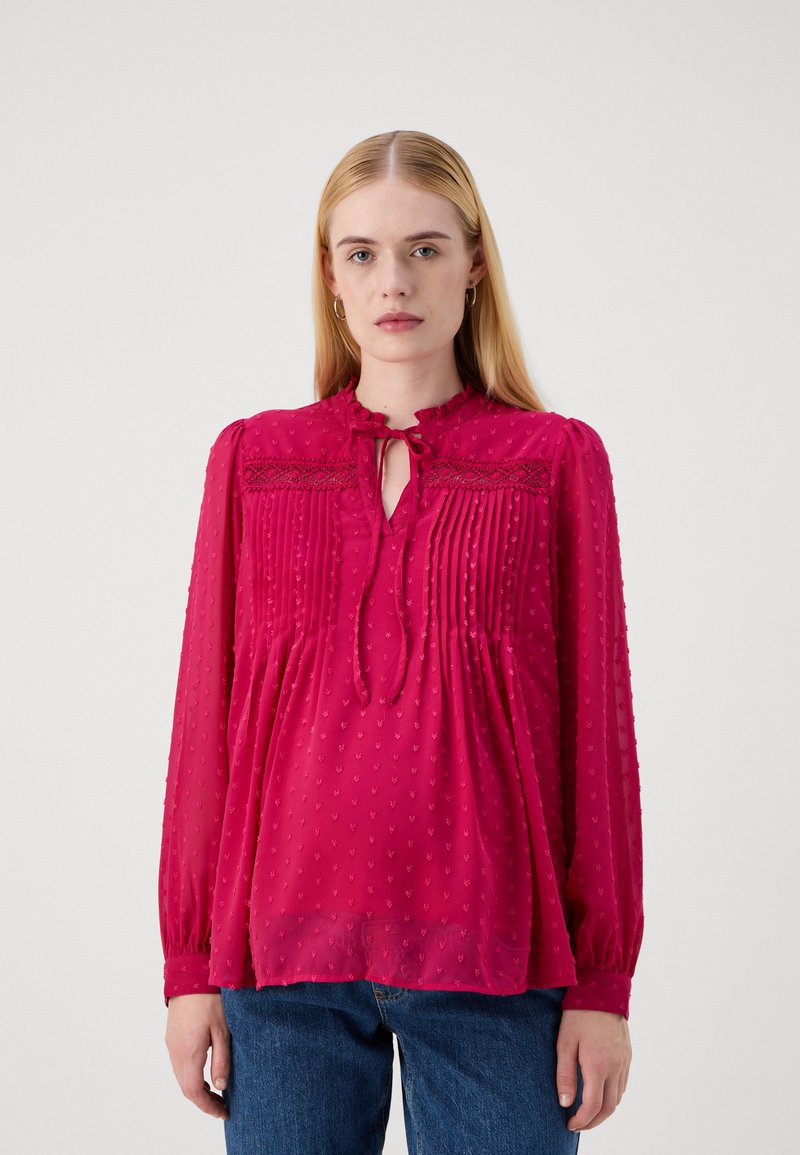 Blusa color lampone con punti testurizzati, dotata di un colletto a legaccio, colletto a volant e parte anteriore plissettata. Maniche lunghe e vestibilità ampia.