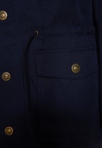 Námořnická modrá bunda vyrobená z látky s hladkou strukturou, s mosaznými knoflíky, šňůrkou v pase a přední kapsou s viditelným šitím.