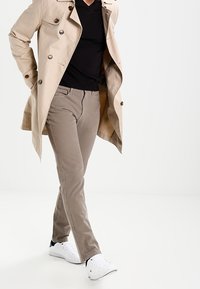 Beige trenchcoat over een zwart V-hals shirt, gecombineerd met lichtbruine broeken en witte sneakers. De jas heeft grote knopen en zakken.