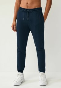 Pantalones deportivos azul marino con cinturilla elástica, cordón, bolsillos laterales y puños acanalados, combinados con zapatillas deportivas blancas.