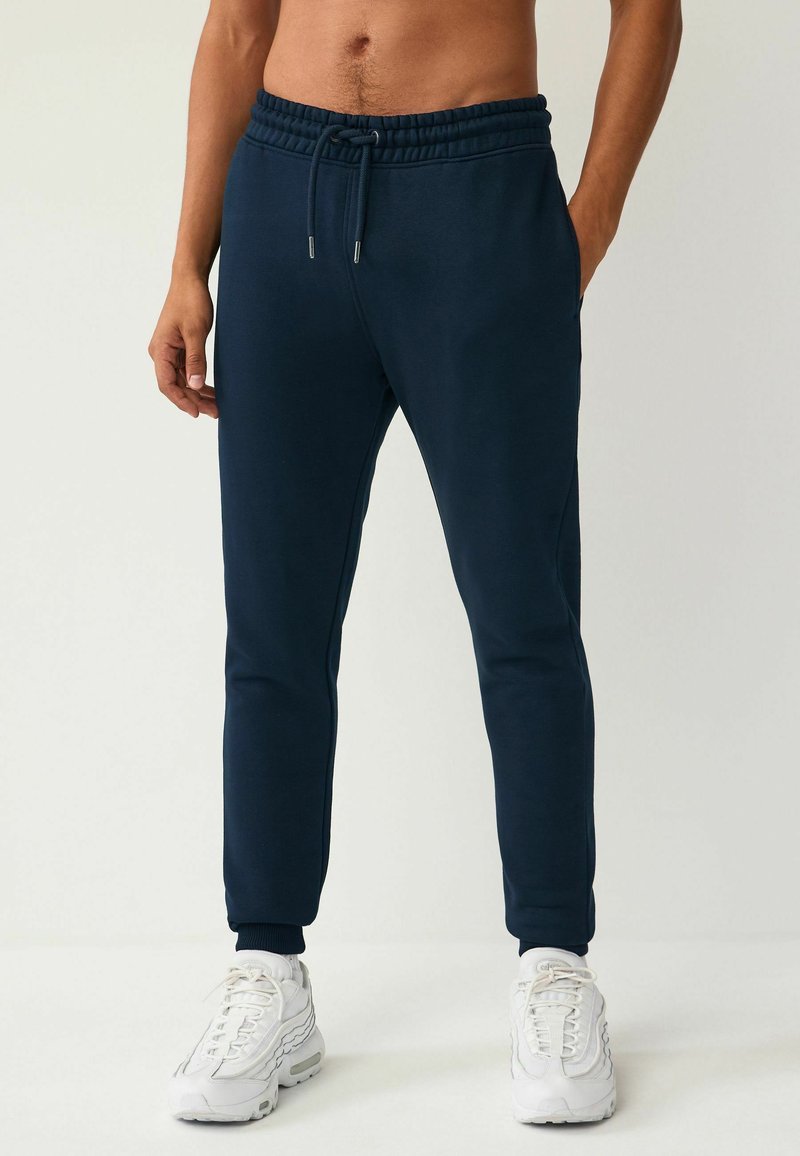 Pantalones deportivos azul marino con cinturilla elástica, cordón, bolsillos laterales y puños acanalados, combinados con zapatillas deportivas blancas.