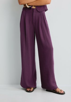 Personne portant un pantalon violet ample taille haute en tissu texturé et des sandales plates noires avec des accents dorés, debout, bras croisés.
