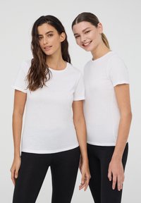 Dwie kobiety noszą dopasowane, krótko rękawowe białe koszulki z bawełny, zestawione z czarnymi legginsami. Koszulki mają okrągły dekolt i klasyczny krój.