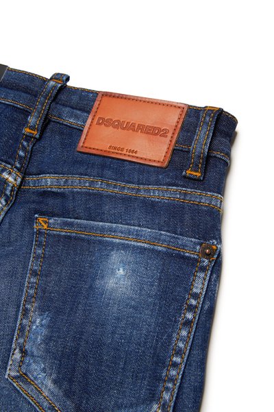 Jeans in denim con un lavaggio blu scuro, caratterizzati da una toppa in pelle marrone con testo impresso, cuciture gialle e dettagli invecchiati sulle tasche.