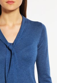 Suéter de punto azul con escote en V y un detalle de nudo en el frente. Mangas largas y una textura suave. Color uniforme sin patrones visibles.