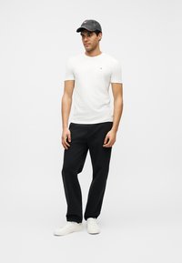 Νεαρός άνδρας με λευκό t-shirt, μαύρο παντελόνι, λευκά αθλητικά παπούτσια και σκούρο καπέλο baseball, στέκεται μπροστά σε ένα απλό φωτεινό φόντο.