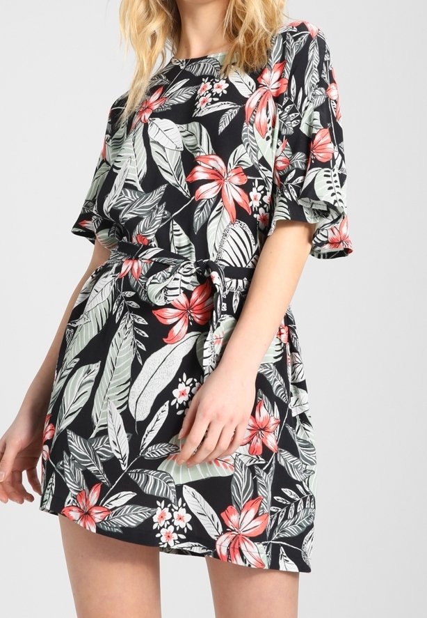 Femme portant une robe noire courte avec un motif floral tropical rouge, blanc et vert, dotée de manches courtes à volants et d'une ceinture à la taille.