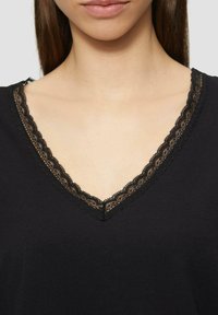 Musthave musta V-kaelusega särk, mis on valmistatud pehmest kangast ning millel on kaeluse ääres detailne must pitsäär, mis täiendab tekstuuri ja visuaalset huvi.