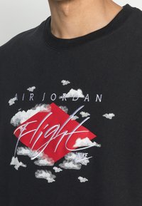 Black cotton t-shirt featuring a red diamond graphic with white clouds. "AIR JORDAN" and "Flight" printed in white above and below, respectively.