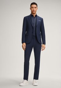 Marineblauw pak van gladde stof, met een getailleerde pasvorm en een colbert met twee knopen, gecombineerd met een bijpassend shirt en witte sneakers.