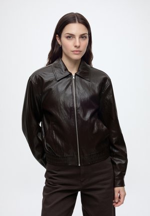 Femme aux longs cheveux foncés, portant une veste en cuir brun foncé zippée et un pantalon de couleur sombre, debout devant un fond clair uni.