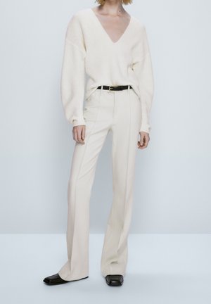 Pull en maille côtelée ivoire avec un décolleté en V profond, associé à un pantalon ajusté couleur crème et une ceinture noire. Chaussures à bout carré noires.