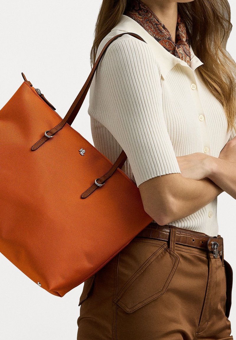 Sac fourre-tout orange avec une finition texturée, poignées en cuir marron et un petit logo. Porté avec un haut crème à côtes et un pantalon marron.