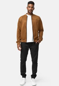 Chaqueta bomber de ante marrón con cremallera y detalles en negro, combinada con pantalones negros ajustados y zapatillas blancas, contra un fondo liso.