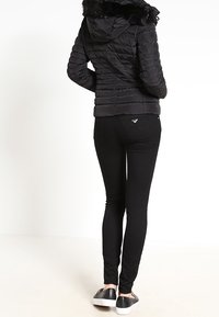 Žena oblečená v černé péřové bundě s kožíškem, černých úzkých džínech a černých slip-on botách, stojící zády k fotoaparátu.