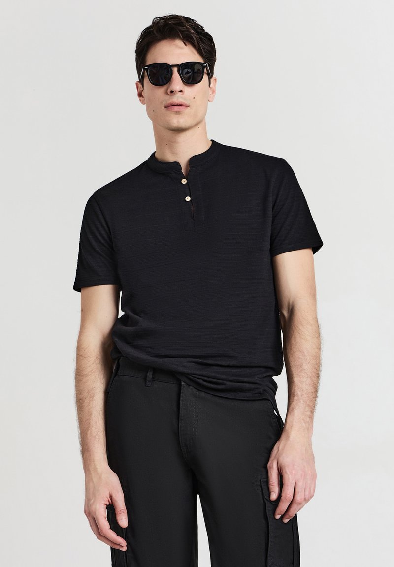 Maglietta henley nera a maniche corte in tessuto texturizzato con tre bottoni in legno sul davanti; abbinata a pantaloni cargo neri e occhiali da sole.