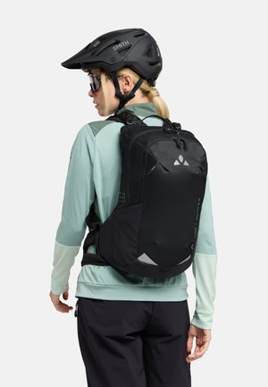 Schwarzer Rucksack mit sleekem Design, strukturiertem Stoff und reflektierenden Akzenten. Getragen über einem hellgrünen Langarmoberteil mit dunkler Hose.