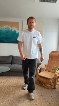 T-shirt branco da Levi's com logótipo no peito esquerdo, jeans pretos e ténis cinzentos; a caminhar numa sala com um sofá cinzento e uma cadeira de baloiço.