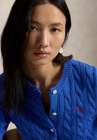 Mlada ženska z dolgimi temnimi lasmi nosi modro jopico iz kvačkanega blaga z belimi gumbi in uhančki v obliki školjk na enobarvnem ozadju.