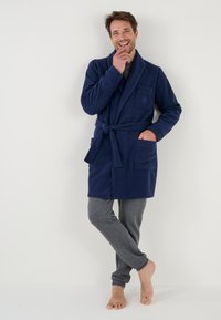 Marineblaue Fleece-robe mit Schalkragen, Taillengurt und Seitentaschen, kombiniert mit grauen Sweatpants und baren Füßen. Weiche Textur.