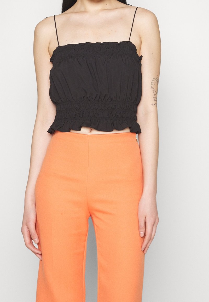Femme portant un top court froncé noir avec fines bretelles et un pantalon orange taille haute, debout sur un fond blanc.