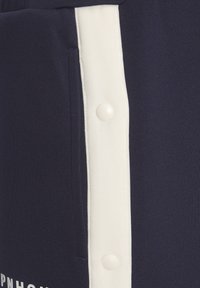 Pantalones cortos deportivos de color azul marino con una franaja lateral blanca y botones a presión, fabricados en un tejido suave y elástico. Bolsillo plano visible.