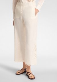 Pantaloni larghi in lino di colore crema chiaro, con intricati dettagli a taglio nel fondo e tasche laterali a soffietto per una maggiore comodità.