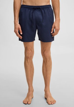 Man draagt knielange marineblauwe gestructureerde korte broek met elastische tailleband en trekkoord, staat blootsvoets tegen een effen achtergrond.