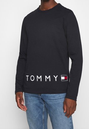 Mężczyzna noszący czarną koszulę z długim rękawem z napisem „TOMMY” i czerwoną, białą oraz czarną prostokątną grafiką na dolnej przedniej części, w połączeniu z niebieskimi dżinsami.