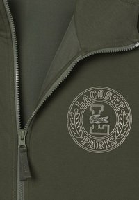 Giacca zip-up verde oliva con tessuto texturizzato, caratterizzata da un logo ricamato e dettagli accentuati in un design circolare.