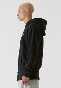 Svart huvtröja i bomull, med en frontficka, ribbade ärmslut och en avslappnad passform. Modellen visas från sidan.