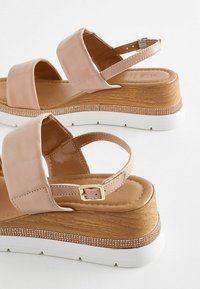 Light pink leather sandals with wide straps, a wooden wedge heel, white rubber sole, and small metallic accents along the base.